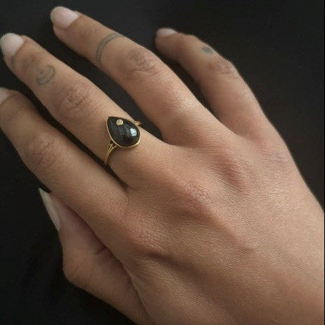 A close-up of a hand wearing a gold ring with a teardrop-shaped black stone, set against a dark background.