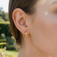A close-up of a woman's ear wearing a small gold hoop earring with three parallel horizontal bands, set against a softly blurred garden background.