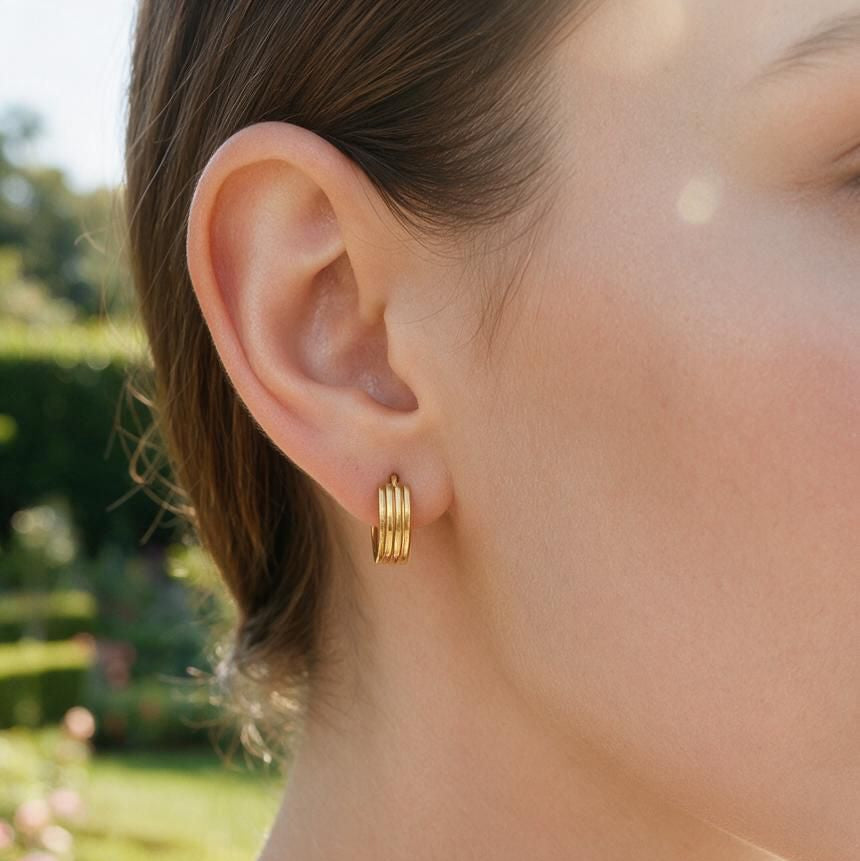 A close-up of a woman's ear wearing a small gold hoop earring with three parallel horizontal bands, set against a softly blurred garden background.